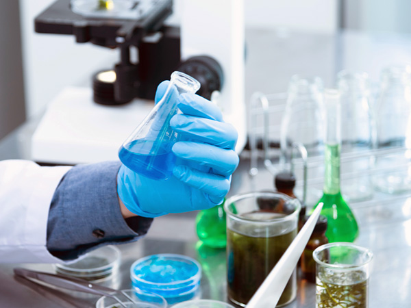 A tabletop covered with laboratory equipment. In the foreground, a hand in a latex glove holds a flask with a blue liquid