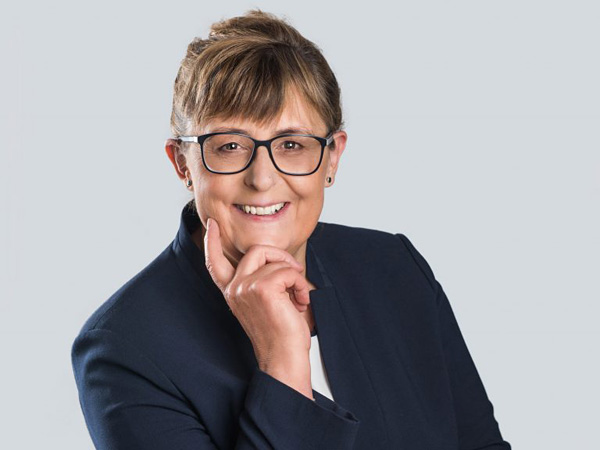 A woman in a dark suit, glasses and her hair tied back. She supports her head with her hand and smiles at the camera