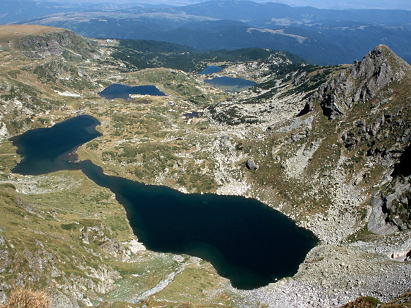 Aerial view of a group of several ponds surrounded by rocky mountains. In the distance, lower mountains covered with forest